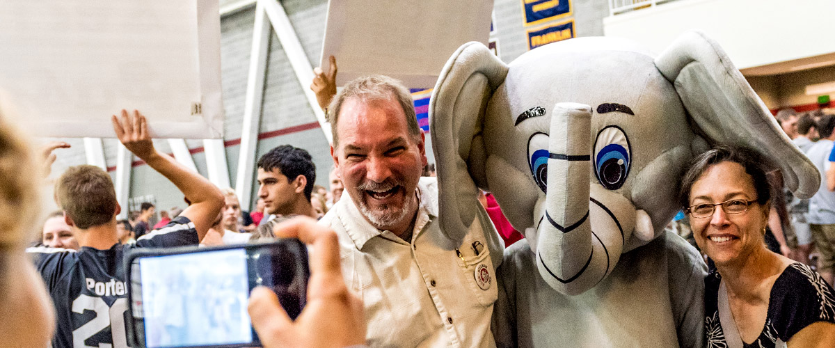 Image shows a student shooting a photo of her smiling parents standing arm-in-arm with Rosie, the 山Ƶ elephant mascot. They are inside a busy Sports and Recreation Center.